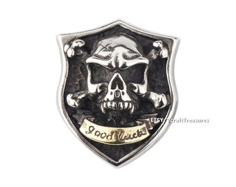 May include: A silver shield pendant with a skull and crossbones design. The skull has a detailed face with sharp teeth and the words "Good Luck" are inscribed on a banner below the skull.