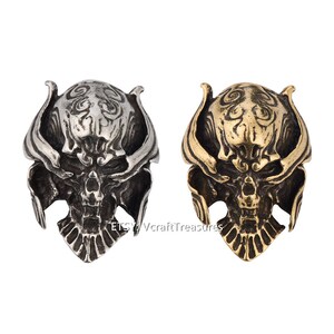 Puede incluir: Dos abalorios de metal con forma de calavera ornamentada, uno plateado y otro dorado, con detalles intrincados y una expresión amenazante.