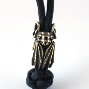 May include: A brass bat charm with a detailed design, hanging from a black cord. The bat has a menacing expression with sharp teeth and wings.