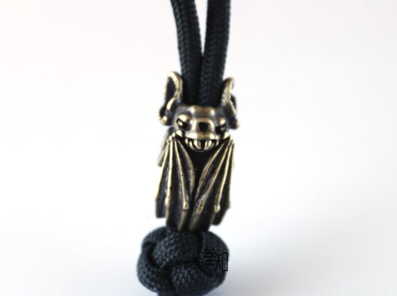 May include: A brass bat charm with a detailed design, hanging from a black cord. The bat has a menacing expression with sharp teeth and wings.