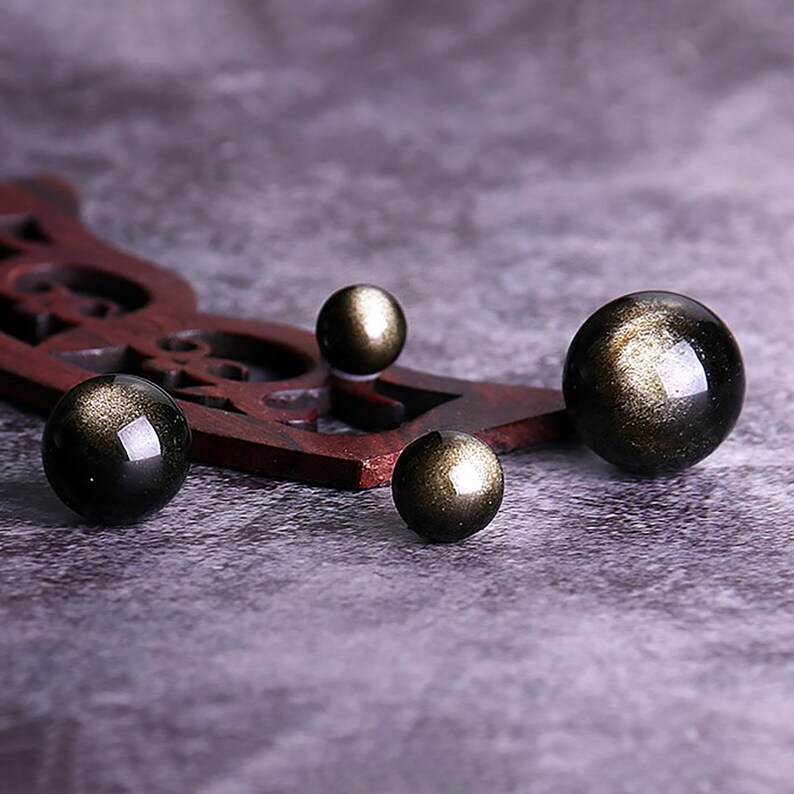 K&ouml;nnte beinhalten: Vier schwarze Obsidiankugeln mit goldenen Sprenkeln, angeordnet auf einer Holzoberfl&auml;che. Die Kugeln sind in verschiedenen Gr&ouml;&szlig;en.