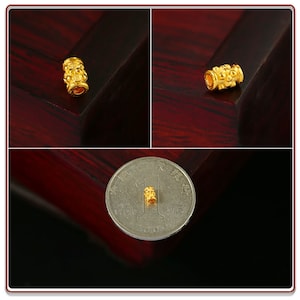 May include: A gold-colored cylindrical bead with intricate designs. The bead is small and is shown next to a Chinese yuan coin for scale.