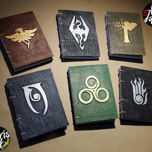 Puede incluir: Seis diarios rectangulares con cubiertas texturizadas en marrón, negro, dorado, verde y gris oscuro. Cada cuaderno presenta un emblema metálico distintivo en la parte frontal, incluyendo un dragón, un árbol y varios símbolos, encuadernados con costura de cordel visible.