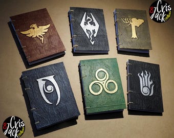 May include: Six rectangular journals with textured covers in brown, black, gold, green, and dark gray. Each notebook features a distinct metallic emblem on the front, including a dragon, a tree, and various symbols, bound with visible cord stitching.