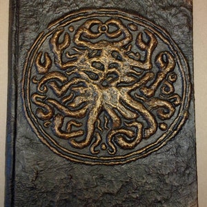 Puede incluir: Un diario de cuero marr&oacute;n oscuro con un dise&ntilde;o en relieve dorado de una criatura con tent&aacute;culos en la portada.