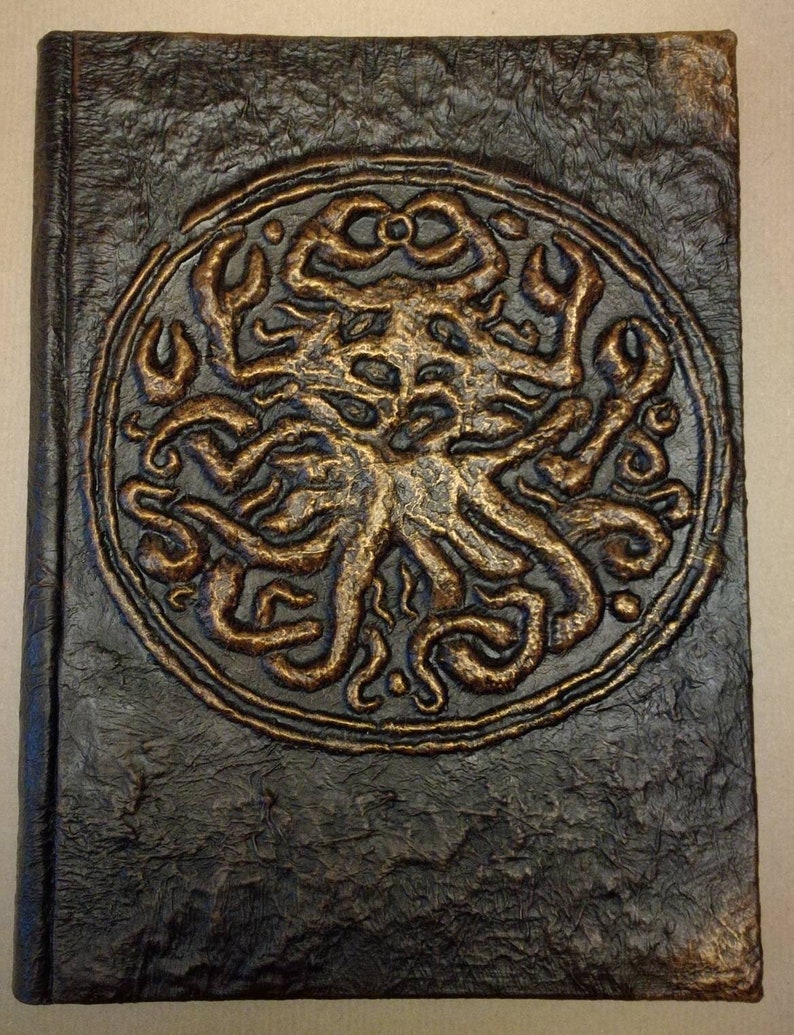 Puede incluir: Un diario de cuero marr&oacute;n oscuro con un dise&ntilde;o en relieve dorado de una criatura con tent&aacute;culos en la portada.