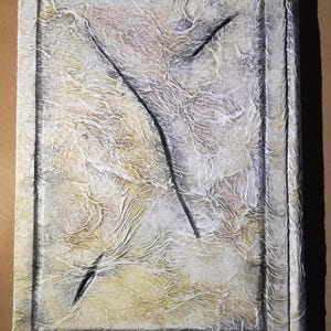 Op de afbeelding: Getextureerd rechthoekig kunstwerk met een cr&egrave;me- en gele achtergrond, omlijst door een donkergrijze rand. Een zwart, abstract ontwerp verdeelt het midden. Het oppervlak heeft een gerimpeld, gelaagd uiterlijk.