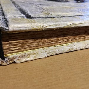 Op de afbeelding: Close-up van een handgemaakt boek met verouderde, getextureerde pagina's. De kaft is een licht, getextureerd materiaal met een verweerde uitstraling en de rug is afgerond. De randen van de pagina's zijn gevlekt, wat het een vintage look geeft.