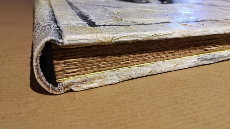 Op de afbeelding: Close-up van een handgemaakt boek met verouderde, getextureerde pagina's. De kaft is een licht, getextureerd materiaal met een verweerde uitstraling en de rug is afgerond. De randen van de pagina's zijn gevlekt, wat het een vintage look geeft.
