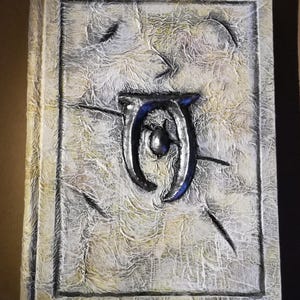 Op de afbeelding: Een getextureerd, gebroken wit boek met een verhoogd, zilverkleurig embleem met blauwe accenten. De kaft heeft een verweerde uitstraling met een rechthoekige rand en een getextureerd oppervlak. De rug van het boek is zichtbaar aan de linkerkant.