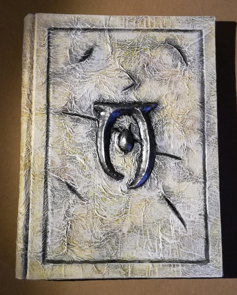 Op de afbeelding: Een getextureerd, gebroken wit boek met een verhoogd, zilverkleurig embleem met blauwe accenten. De kaft heeft een verweerde uitstraling met een rechthoekige rand en een getextureerd oppervlak. De rug van het boek is zichtbaar aan de linkerkant.