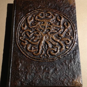 Puede incluir: Un diario encuadernado en cuero marr&oacute;n con un dise&ntilde;o circular en relieve de una criatura con tent&aacute;culos de color cobre.