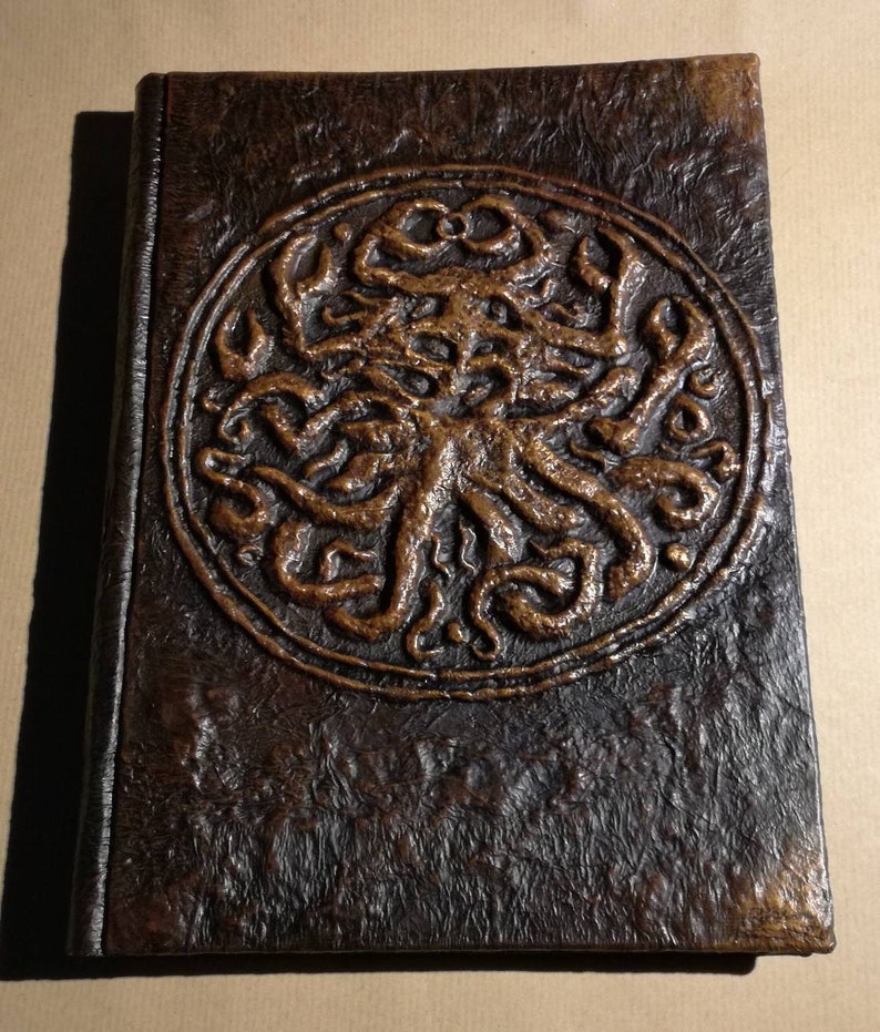 Puede incluir: Un diario encuadernado en cuero marr&oacute;n con un dise&ntilde;o circular en relieve de una criatura con tent&aacute;culos de color cobre.