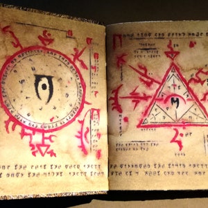 Op de afbeelding: Open boek met verouderde papieren pagina's met rode en zwarte symbolen en tekst. E&eacute;n pagina heeft een cirkelvormig ontwerp met een centraal oogsymbool. De andere pagina heeft een driehoek met een centraal symbool. Het boek heeft een gouden rand.
