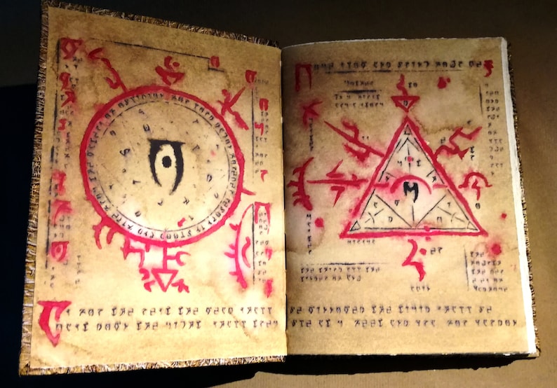Op de afbeelding: Open boek met verouderde papieren pagina's met rode en zwarte symbolen en tekst. E&eacute;n pagina heeft een cirkelvormig ontwerp met een centraal oogsymbool. De andere pagina heeft een driehoek met een centraal symbool. Het boek heeft een gouden rand.