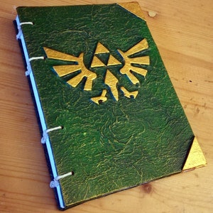 Puede incluir: Un diario encuadernado en cuero verde con un símbolo Triforce dorado en relieve en la portada.