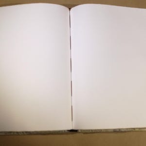 Op de afbeelding: Een open boek met blanco witte pagina's. Het boek heeft een goudkleurige rand op de kaft en randen. Het boek ligt open op een bruin oppervlak, klaar om te schrijven of te tekenen.