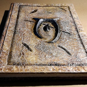Op de afbeelding: Een handgemaakt, getextureerd boek met een verweerde uitstraling. De kaft heeft een verhoogd, donker metalen embleem en decoratieve ge&euml;tste lijnen. De randen van het boek zijn ook getextureerd, wat het een vintage esthetiek geeft. Het kleurenpalet omvat tinten bruin, beige en grijs.