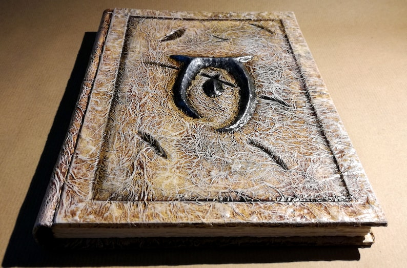 Op de afbeelding: Een handgemaakt, getextureerd boek met een verweerde uitstraling. De kaft heeft een verhoogd, donker metalen embleem en decoratieve ge&euml;tste lijnen. De randen van het boek zijn ook getextureerd, wat het een vintage esthetiek geeft. Het kleurenpalet omvat tinten bruin, beige en grijs.
