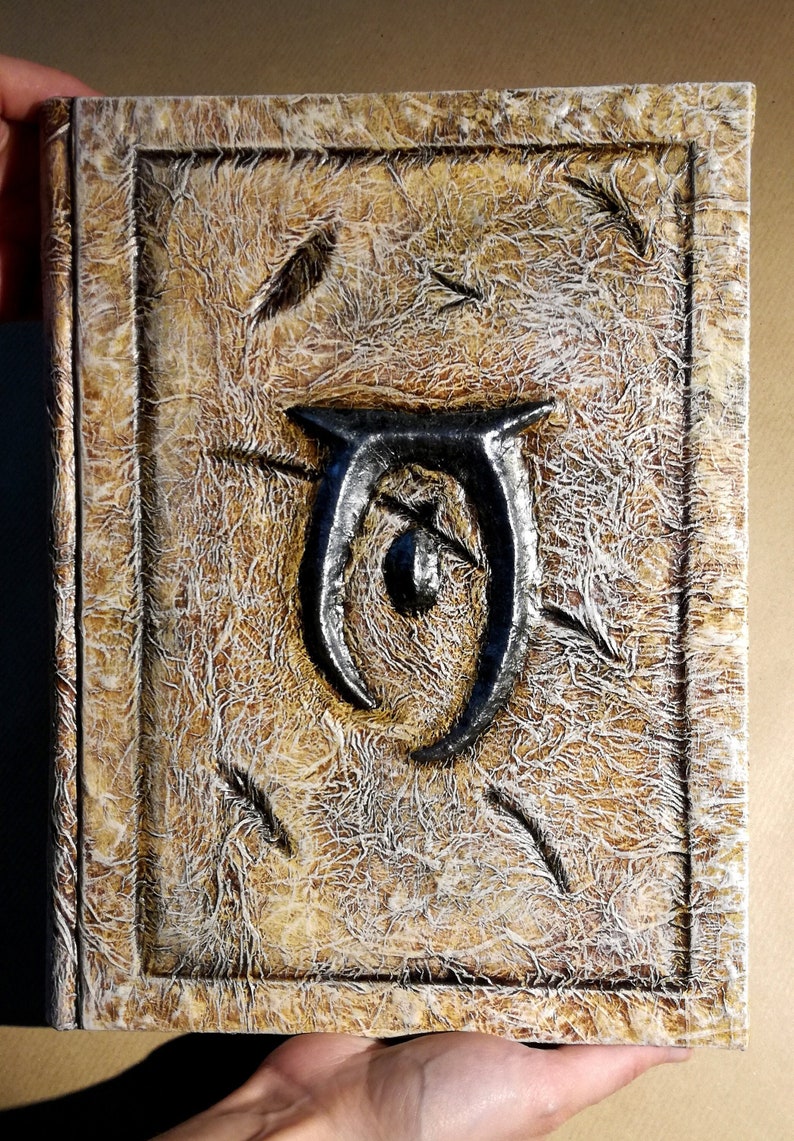 Op de afbeelding: Een handgemaakt boek met een getextureerde, verouderde uitstraling. De kaft heeft een verheven, metalen embleem en decoratieve, veerachtige accenten. Het kleurenpalet omvat tinten bruin, beige en wit, wat een vintage esthetiek cre&euml;ert. Het boek wordt door een persoon vastgehouden.