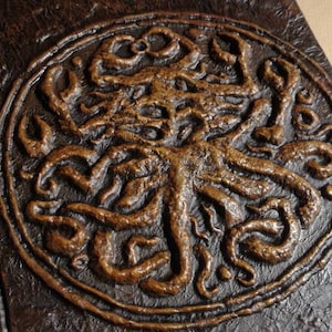 Puede incluir: Un diario encuadernado en cuero marr&oacute;n con un dise&ntilde;o circular en relieve de una criatura con tent&aacute;culos de color cobre.