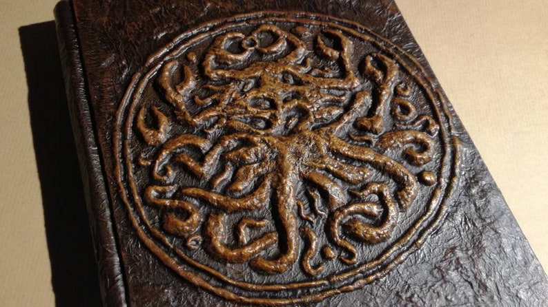 Puede incluir: Un diario encuadernado en cuero marr&oacute;n con un dise&ntilde;o circular en relieve de una criatura con tent&aacute;culos de color cobre.