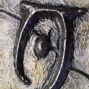 Op de afbeelding: Close-up van een gestructureerd kunstwerk met een donker, metalen symbool tegen een achtergrond van gelaagde, gebroken witte en gele tinten. Het symbool is een gestileerd, gebogen ontwerp met een centrale bol.
