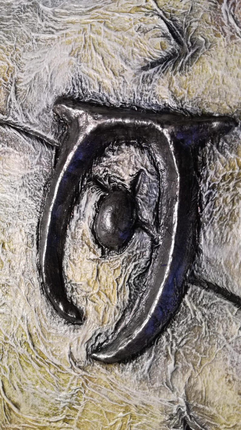 Op de afbeelding: Close-up van een gestructureerd kunstwerk met een donker, metalen symbool tegen een achtergrond van gelaagde, gebroken witte en gele tinten. Het symbool is een gestileerd, gebogen ontwerp met een centrale bol.
