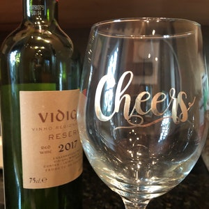 May include: A glass wine glass with the word "Cheers" etched in gold on the side. A bottle of red wine is in the background.