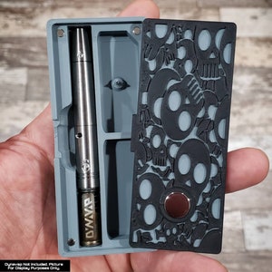 May include: A gray and black storage case with a skull pattern on the lid. The case has a compartment for a vape pen and a small compartment for a battery. The case is made of plastic and has a magnetic closure.