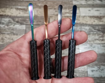 May include: Four different colored metal tools with carbon fiber handles. The tools have a flat, rounded tip and are used for scraping or spreading.