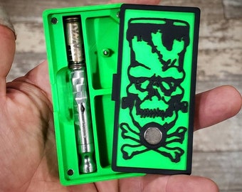 Peut inclure: Une boîte de rangement verte et noire avec un motif de crâne et d'os croisés. La boîte a un compartiment pour un stylo vape Dynavap. Dynavap non inclus. Image à des fins d'illustration uniquement.