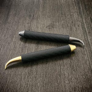 May include: Two black handled tools with curved blades. One has a silver blade, the other a gold blade. The tools are lying on a dark wood surface.