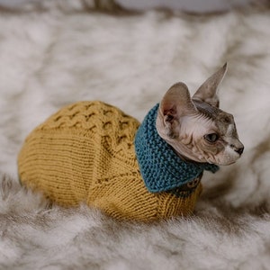 May include: A Sphynx cat wearing a mustard yellow knitted sweater with a diamond pattern and a teal blue collar. The cat is sitting on a fluffy, white faux fur blanket.