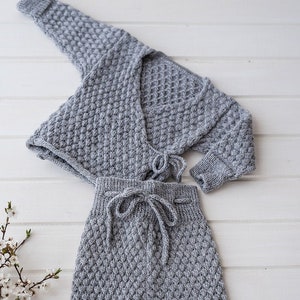 Op de afbeelding: Een grijze gebreide baby-outfit met een lang mouw cardigan en bijpassende broek. De cardigan heeft een wikkel-sluiting en een strik in de taille. De broek heeft een trekkoord in de taille en een ontspannen pasvorm.