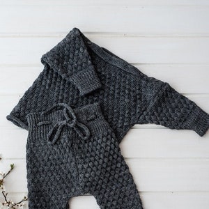 Op de afbeelding: Een grijze gebreide baby-outfit met een vest en broek. De outfit heeft een textuur, honingraat-achtig patroon.