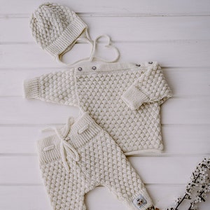 Op de afbeelding: Een witte gebreide baby-outfitset bestaande uit een muts, trui en broek. De set is gemaakt van zacht, getextureerd garen en heeft een eenvoudig, klassiek ontwerp.