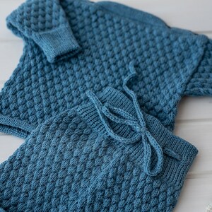 Op de afbeelding: Een blauwe gebreide babytrui en broekset met een textuurpatroon. De set is gemaakt van zacht garen en heeft een comfortabele pasvorm.