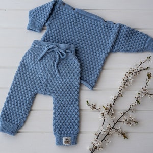 Op de afbeelding: Een blauwe gebreide babytrui en broekset met een textuurpatroon. De set ligt op een wit houten oppervlak met een tak met witte bloesems.