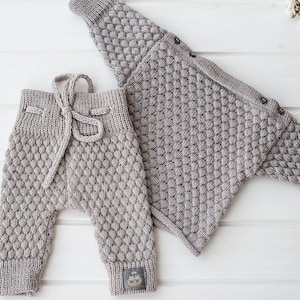 Op de afbeelding: Een grijze gebreide babytrui en broek set met een textuur, bobbelsteek patroon. De trui heeft een knoopsluiting aan de hals.