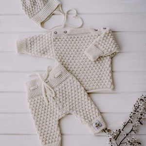 Op de afbeelding: Een witte gebreide baby outfit set bestaande uit een muts, trui en broek. De set is gemaakt van zacht, getextureerd garen en heeft een eenvoudig, klassiek ontwerp.
