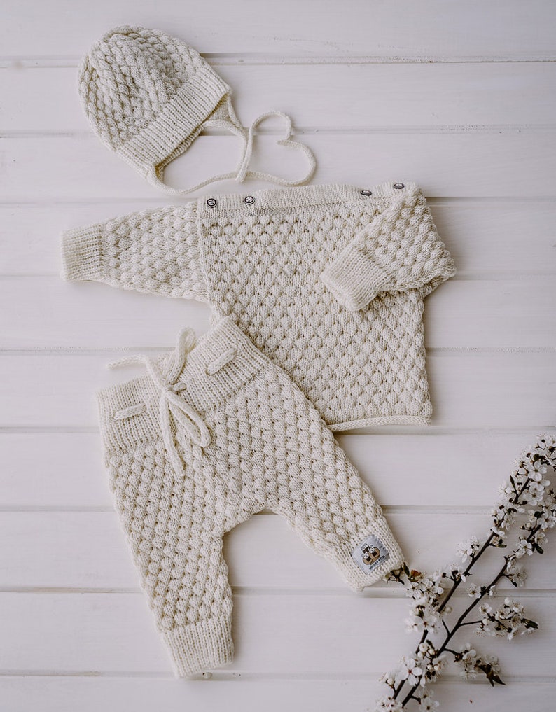 Op de afbeelding: Een witte gebreide baby outfit set bestaande uit een muts, trui en broek. De set is gemaakt van zacht, getextureerd garen en heeft een eenvoudig, klassiek ontwerp.