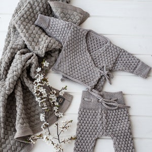 Op de afbeelding: Een grijze gebreide baby-outfitset inclusief een cardigan, broek en een deken. De cardigan heeft een wikkel-stijl met een strik sluiting. De broek heeft een trekkoord tailleband. De deken is textuur en heeft een wafel-achtig patroon.