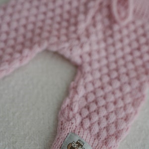 Op de afbeelding: Roze gebreide babybroek met een geweven label met de tekst "Woolen Wool" en een cartoonafbeelding van twee katten.