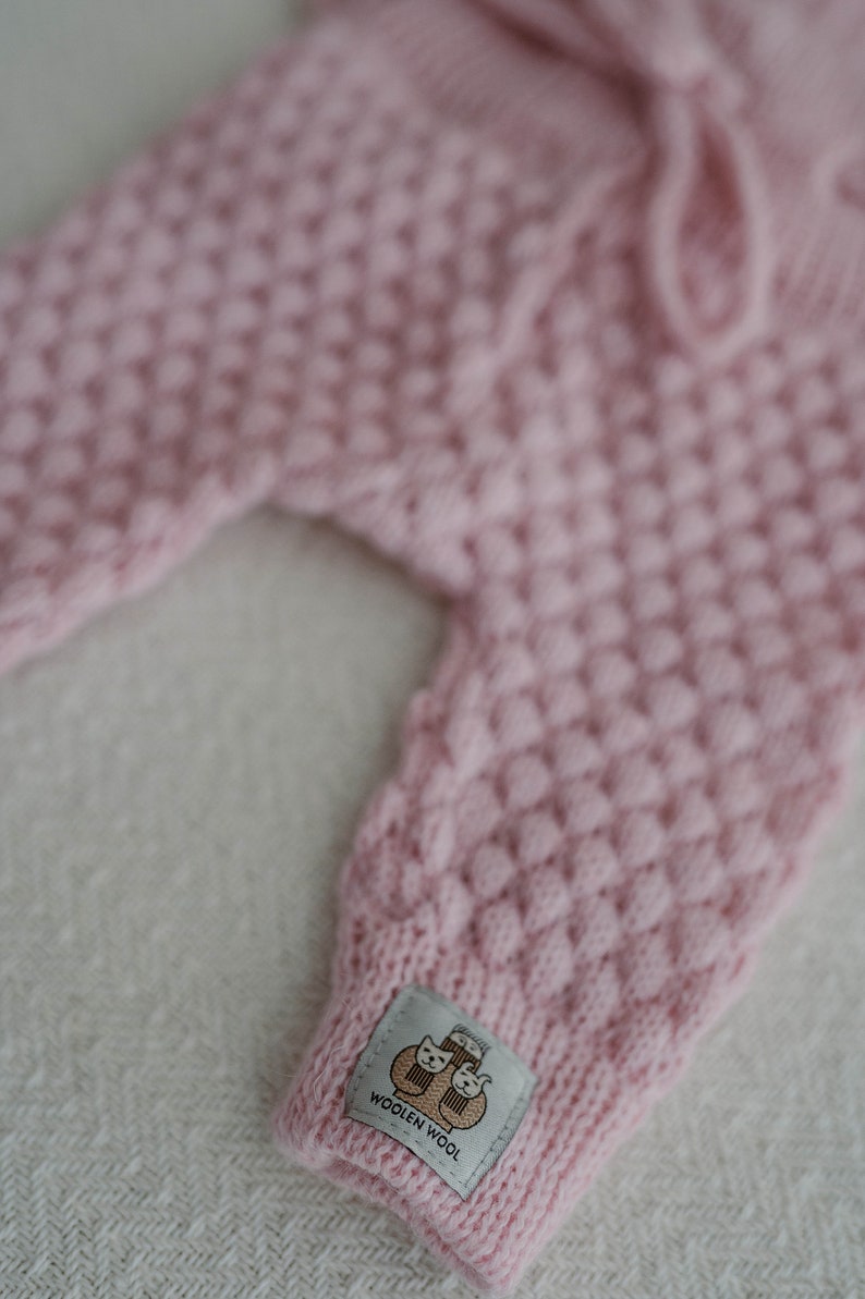 Op de afbeelding: Roze gebreide babybroek met een geweven label met de tekst "Woolen Wool" en een cartoonafbeelding van twee katten.