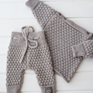 Op de afbeelding: Een gebreide baby-outfit in lichtgrijs. De outfit bestaat uit een vest met knopen en een bijpassende broek met een strik in de taille.