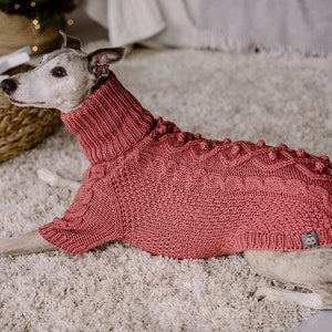 May include: A Whippet dog wearing a pink knitted sweater with a cable knit pattern and pom-poms. The sweater has a turtleneck collar.