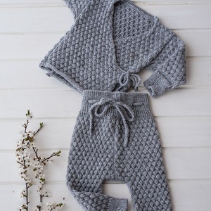 Op de afbeelding: Een grijze gebreide baby-outfit met een cardigan en broek. De cardigan heeft een wikkel-stijl en knoopt in de taille. De broek heeft een trekkoord.