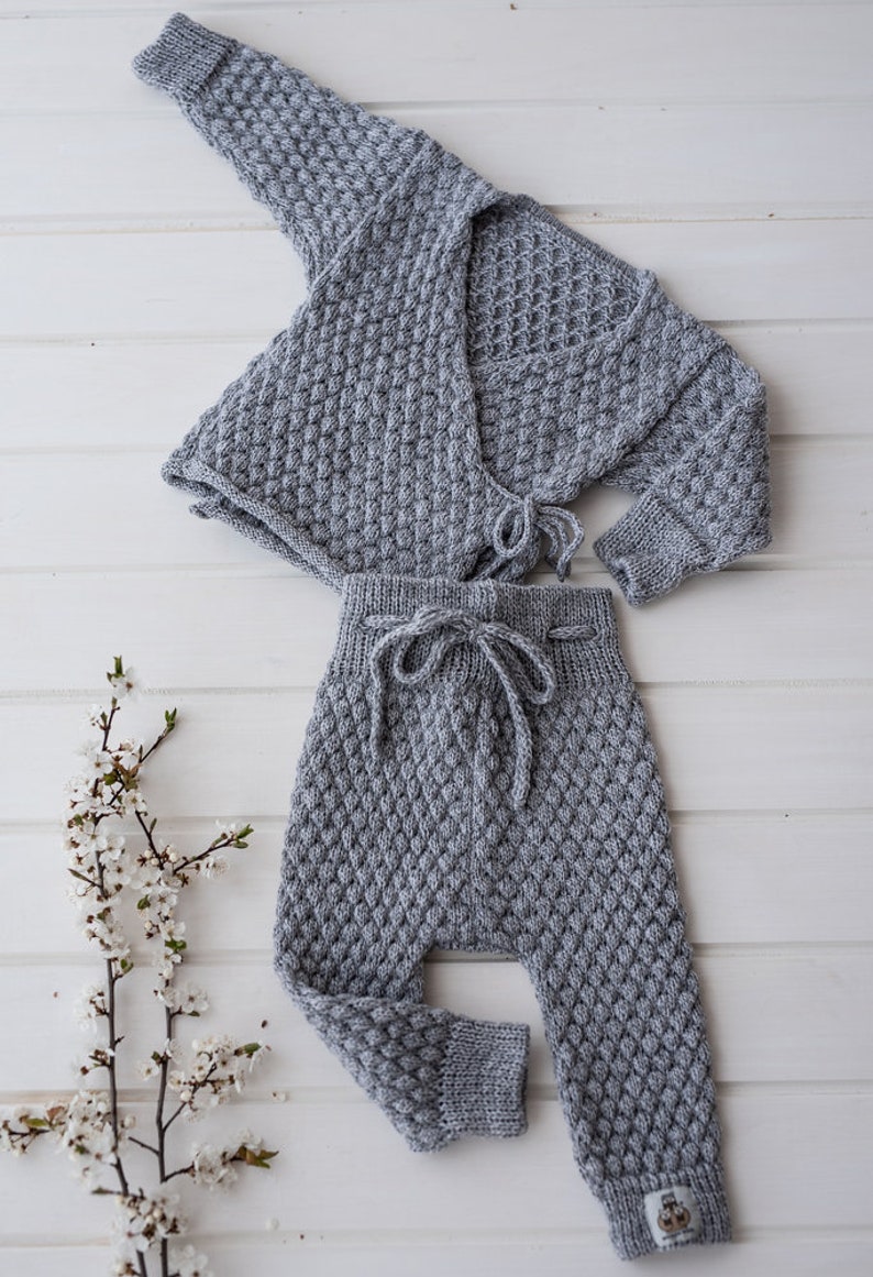 Op de afbeelding: Een grijze gebreide baby-outfit met een cardigan en broek. De cardigan heeft een wikkel-stijl en knoopt in de taille. De broek heeft een trekkoord.