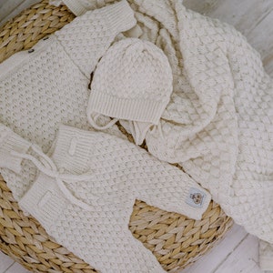 Op de afbeelding: Een witte gebreide baby-outfitset inclusief een muts, een trui en een broek. De set is gemaakt van zacht, getextureerd garen en heeft een delicaat kabelbreipatroon.